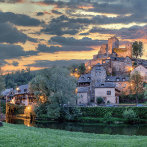 Vue du village de Belcastel en Aveyron au coucher du soleil, avec son château illuminé et la rivière en premier plan.